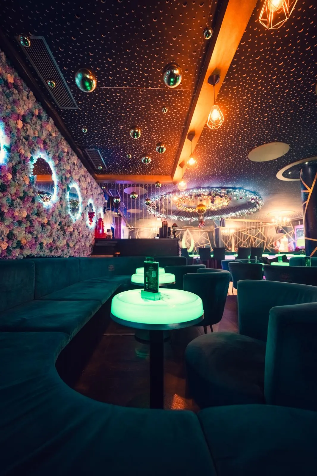 Lounge mit Blumenwand, runden Spiegeln, Samtsofas, leuchtenden Tischen und Discokugeln an der Decke
